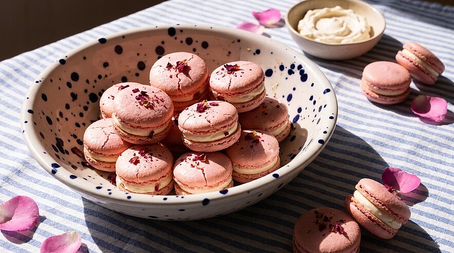 Schüssel mit rosa Macarons, einige mit getrockneten Rosenblättern garniert; weitere Macarons und Rosenblätter liegen neben einer kleinen Schale mit Creme auf gestreiftem Tuch.