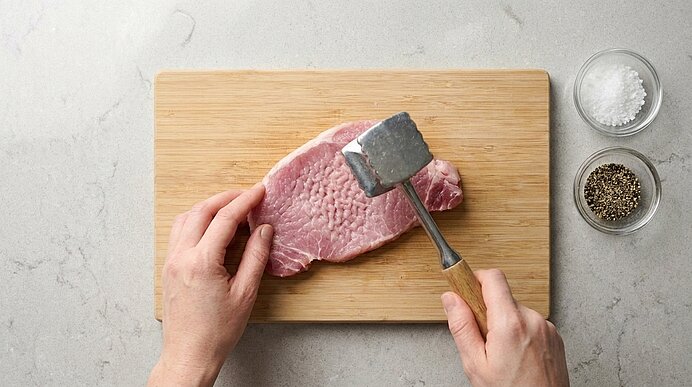 Zwei Hände klopfen ein rohes Schnitzel auf einem Holzbrett mit einem Fleischklopfer. Daneben stehen Salz und Pfeffer.