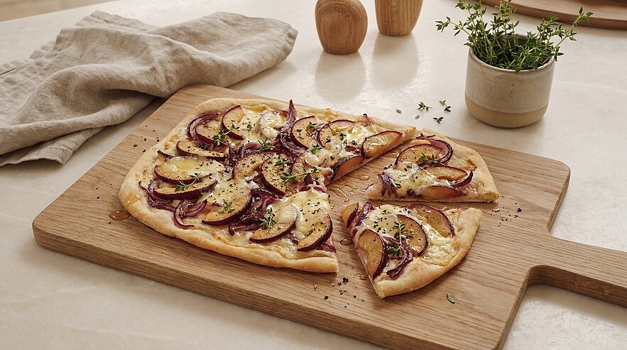 Rechteckiger Flammkuchen auf Holzbrett, belegt mit dünnen Birnenscheiben, roten Zwiebeln, geschmolzenem Käse und frischen Kräutern; ein Stück ist herausgeschnitten.