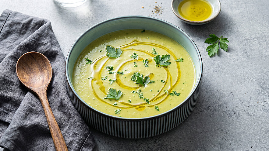 Eine Schüssel mit cremiger Suppe, garniert mit frischen Kräutern und einem Schuss Olivenöl. Neben der Schüssel liegt ein Holzlöffel und ein graues Tuch. Der Hintergrund ist schlicht und unauffällig.