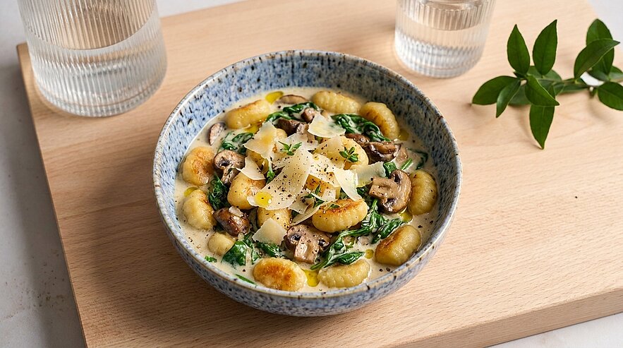 Schüssel mit gebratenen Gnocchi in cremiger Sauce, Champignons, Spinat und Parmesanspänen, mit Kräutern garniert.