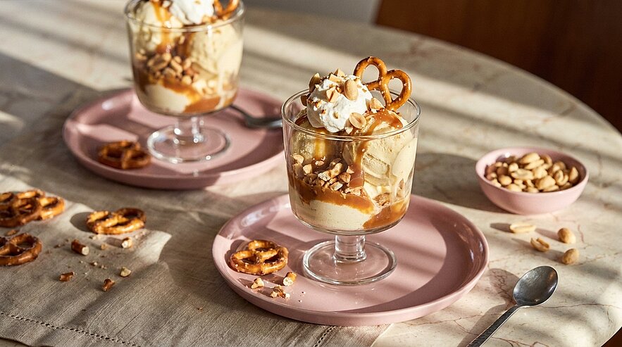 Schichtdessert in Glasbechern mit Karamellsauce, Schlagsahne, salzigen Brezeln und Erdnüssen, serviert auf rosa Tellern.