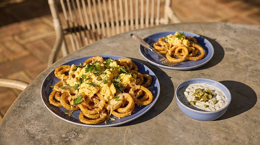 Zwei blaue Teller mit knusprigen, goldbraunen Ringen, obenauf Rührei und frischen Kräutern; daneben eine kleine Schale mit weißer Dip-Sauce und gehackten Gewürzgurken auf einem Tisch.