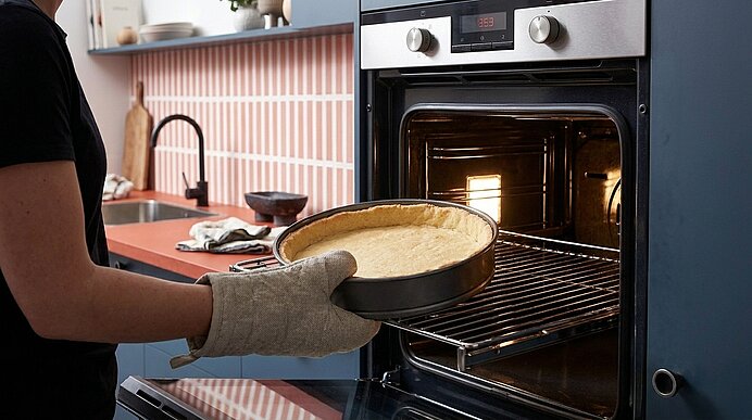 Person mit Ofenhandschuhen schiebt eine runde Kuchenform mit Teig in einen vorgeheizten Backofen.