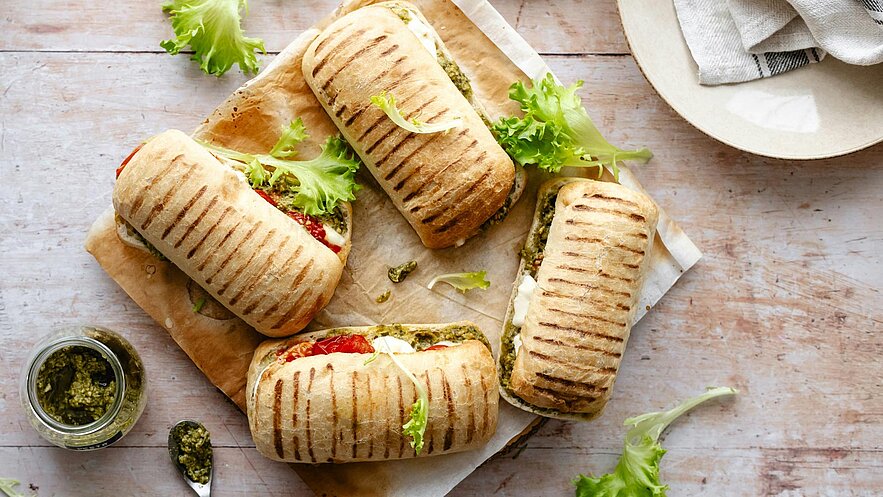 Vier gegrillte Sandwiches mit frischem Gemüse liegen auf einem Holzbrett. Jedes Sandwich ist mit einer grünen Paste und Salat gefüllt. Ein Glas mit grüner Paste und einem Löffel sind ebenfalls in der Nähe. Der Hintergrund ist hell und schlicht.