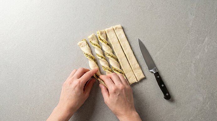 Zwei Hände drehen mit Pesto gefüllte Blätterteigstreifen auf der hellgrauen Arbeitsplatte. Rechts liegt ein Messer.