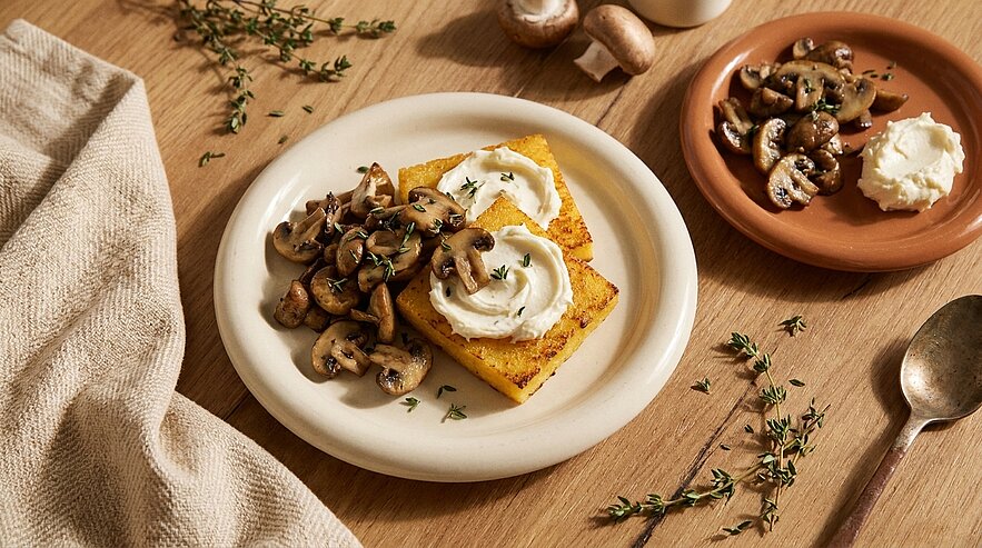 Weißer Teller mit zwei Polenta-Schnitten, belegt mit Frischkäse und sautierten Champignons, mit frischem Thymian garniert.