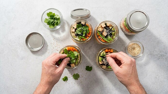 Zwei Hände füllen und garnieren mehrere Einmachgläser mit buntem Salat (Karotten, Gurken, grüne Blätter) mit frischem Koriander und Sesamkörnern auf heller Arbeitsfläche.