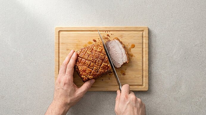 Zwei Hände schneiden mit einem großen Messer einen knusprigen Krustenbraten auf einem Holzbrett; eine Scheibe ist bereits abgeschnitten.