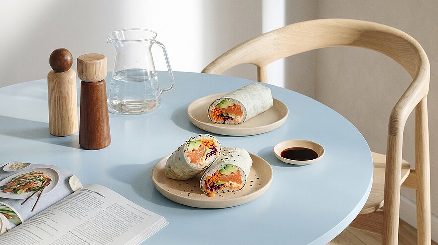Zwei Teller mit Gemüse-Wraps auf hellem blauen Tisch; auf dem Vordergrundteller ein halbierter Wrap mit bunter Gemüsefüllung, daneben eine kleine Schale mit dunkler Sauce.