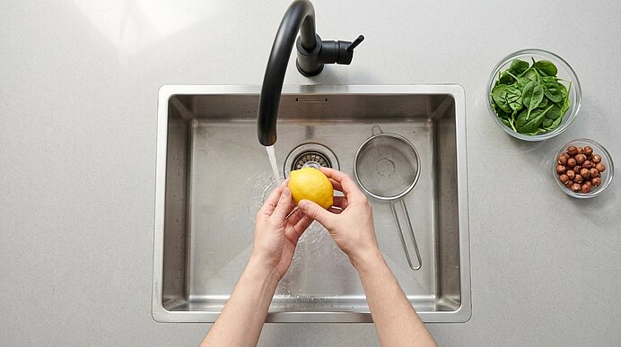 Zwei Hände waschen eine Zitrone unter fließendem Wasser im Edelstahlbecken; rechts stehen Spinat und Haselnüsse.