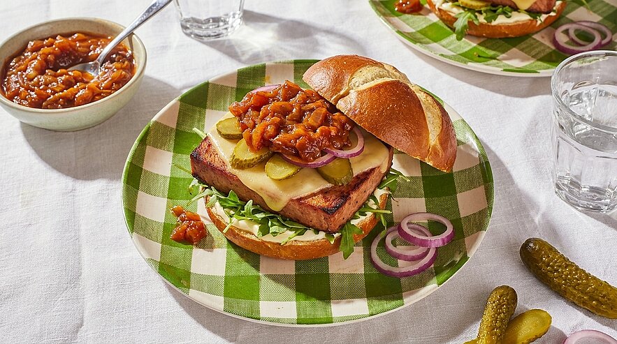 Laugenbrötchen-Burger mit rechteckigem Pflanzenpatty, geschmolzenem Käse, Gewürzgurkenscheiben, roten Zwiebelringen, Salat und würzigem Chutney obenauf.
