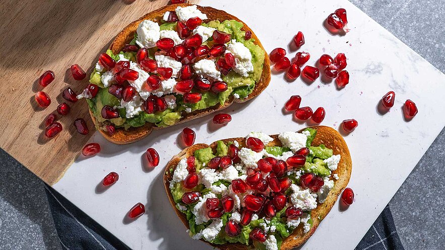 Zwei geröstete Brotscheiben mit einer cremigen Avocado-Aufstrich, garniert mit zerbröckeltem Käse und roten Granatapfelkernen. Der Hintergrund ist eine helle, strukturierte Oberfläche, auf der einige Granatapfelkerne verstreut liegen.