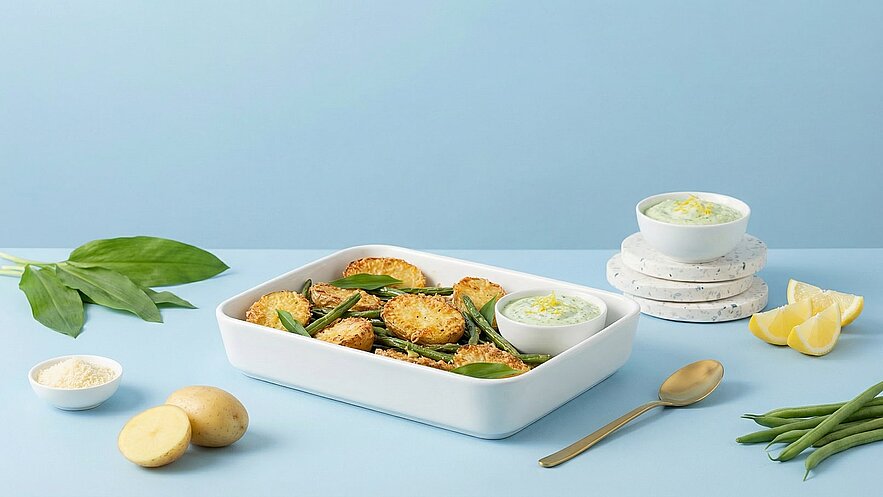Ein weißes Backgericht mit gebackenem Gemüse, darunter grüne Bohnen und knusprige Scheiben. Neben dem Gericht steht eine kleine Schüssel mit Dip und einem goldenen Löffel. Dekorative Zitronenscheiben und frische Kräuter ergänzen die Präsentation. Der Hintergrund ist in sanftem Blau gehalten.