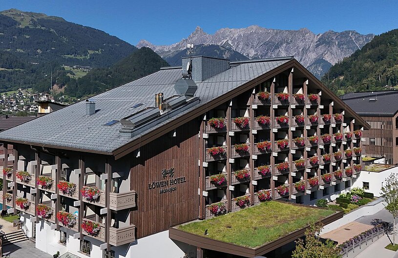 Das Bild zeigt das Löwen Hotel in Mandly, ein modernes Holzgebäude mit Blumenkästen an den Balkonen. Im Hintergrund sind majestätische Berge und ein klarer blauer Himmel zu sehen. Der Bereich vor dem Hotel ist bepflanzt und umfasst einen Pool.