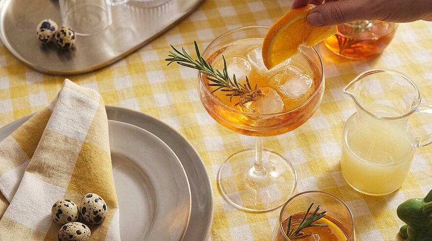 Nahaufnahme einer sonnigen Tischszene: Ein Cocktailglas mit Eis und Rosmarinzweig, eine Hand legt eine Orangenscheibe hinein; daneben ein kleines Glas, ein Milchkännchen und ein Teller mit Stoffserviette und Wachteleiern auf gelb kariertem Tischtuch.