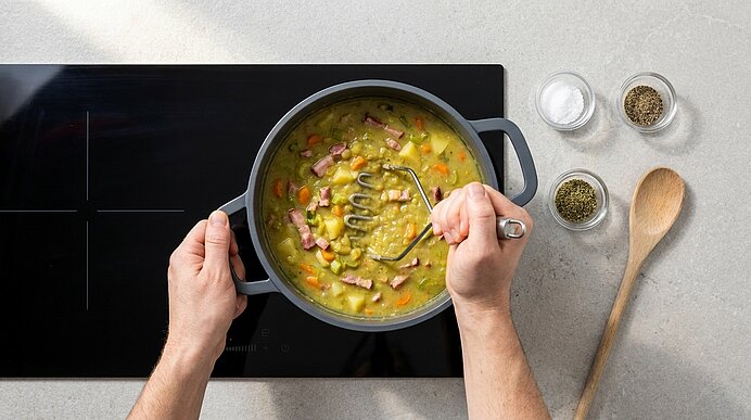 Zwei Hände zerdrücken mit einem Kartoffelstampfer eine dicke Erbsensuppe mit Schinkenwürfeln in einem Topf auf dem Herd.