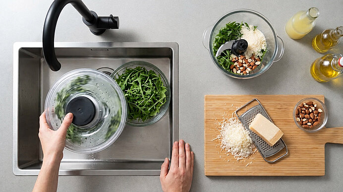 Draufsicht: Rucola wird im Spülbecken geschleudert, Parmesan gerieben und Zutaten im Mixer für Pesto vorbereitet.