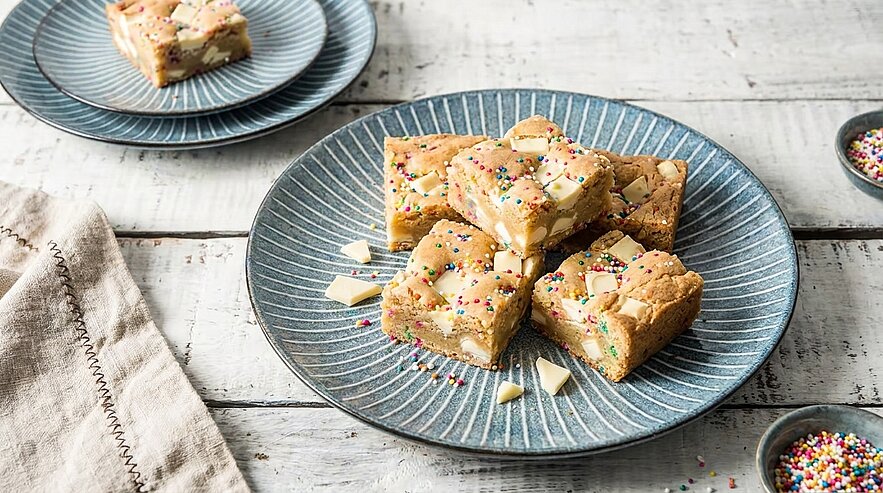 Mehrere eckige Blondie-Stücke mit weißen Schokoladenstücken und bunten Zuckerstreuseln auf einem blauen Keramikteller, auf hellem Holztisch angerichtet.