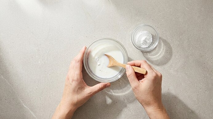 Zwei Hände rühren mit einem Holzlöffel eine weiße Paste in einer Glasschüssel; daneben steht eine kleine Glasschale mit weißem Pulver auf hellem Untergrund.