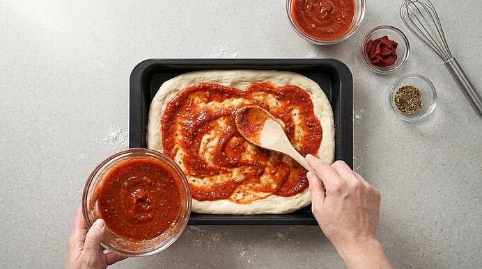 Zwei Hände verteilen mit einem Holzkochlöffel Tomatensauce auf dem Pizzateig in der mattschwarzen Backform.