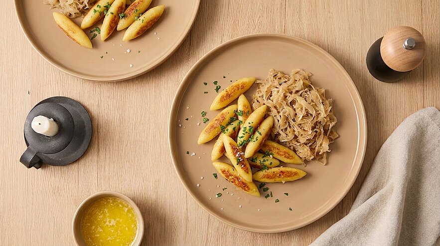 Teller mit goldbraunen, gebratenen Schupfnudeln, bestreut mit gehackten Kräutern, serviert neben einer Portion Sauerkraut.
