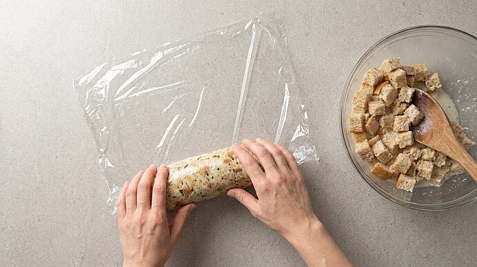 Zwei Hände rollen eine gewürzte Füllungsmasse in Frischhaltefolie zu einer Zylinderform; daneben eine Schüssel mit Brotwürfeln und Holzlöffel.