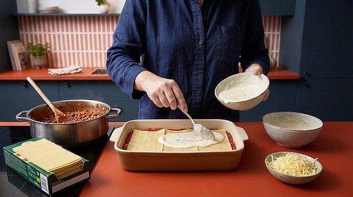 Person verteilt mit Löffel weiße Sauce über eine geschichtete Lasagne in einer Auflaufform; daneben Topf mit Fleischsauce, Stapel Lasagneblätter und Schale mit geriebenem Käse.