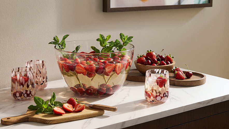 Eine große Schüssel mit Erdbeeren und Minze steht auf einem Tisch. Neben der Schüssel befinden sich zwei Gläser und eine Holzplatte mit weiteren Erdbeeren. Der Tisch hat eine helle Marmoroberfläche.