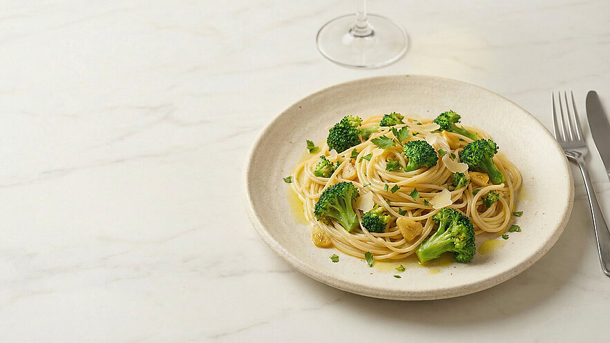 Ein tiefes Teller mit Spaghetti und Brokkoli, garniert mit Petersilie und etwas Parmesan. Der Teller ist auf einem hellen Tisch platziert, im Hintergrund ist ein Glas sichtbar.