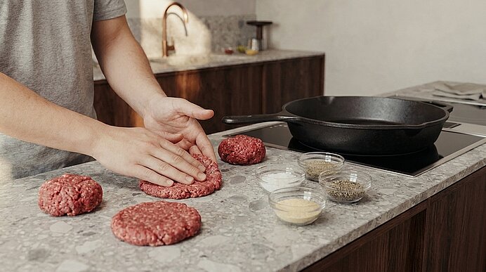 Hände formen rohe Rindfleisch-Patties auf der Küchenarbeitsplatte; daneben eine Pfanne auf dem Herd und kleine Schalen mit Gewürzen.