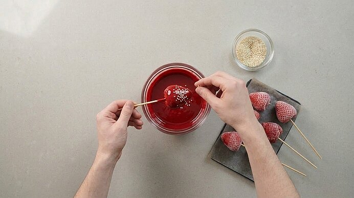 Zwei Hände bestreuen ein rot glasiertes Erdbeer-Dessert am Holzspieß über einer Schüssel roter Kuvertüre mit Sesam.