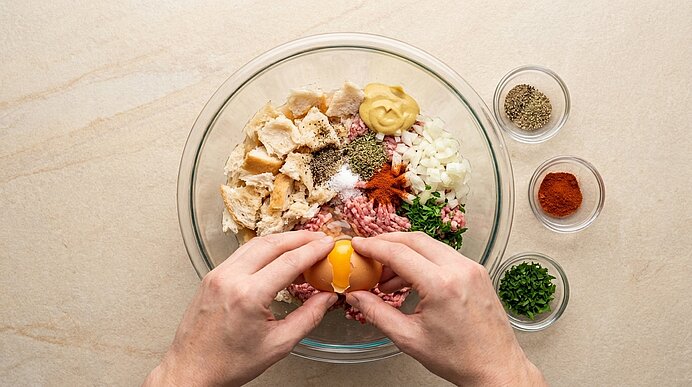 Hände schlagen ein Ei über einer Glasschüssel mit gehacktem Hähnchen, Hackfleisch, Senf, Zwiebeln, Kräutern und Gewürzen; Schälchen mit Pfeffer, Paprika und Schnittlauch daneben.