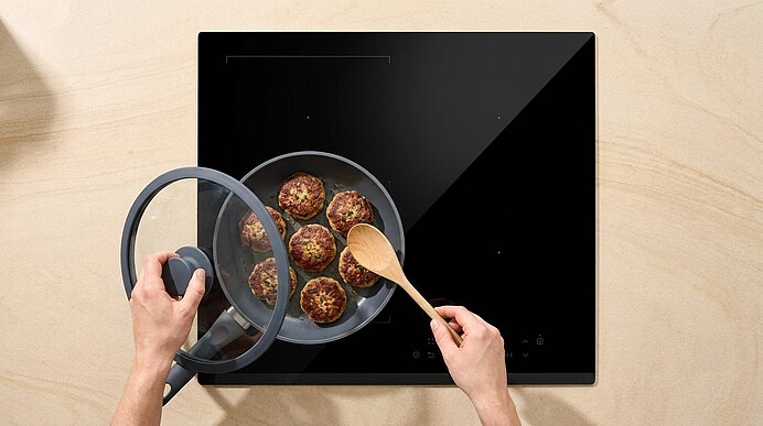 Draufsicht: Hände heben den Glasdeckel einer Pfanne auf einem schwarzen Induktionskochfeld; in der Pfanne werden braun gebratene Fleischbällchen mit einem Holzspatel gewendet.