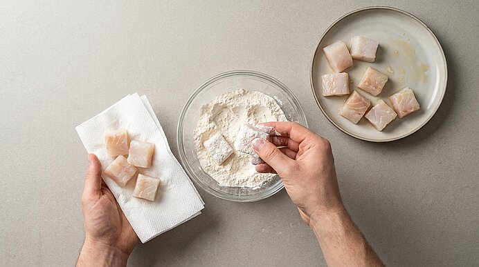 Zwei Hände panieren Fischwürfel: rechte taucht ein Stück in Mehl in einer Glasschüssel, linke hält bemehlte Stücke auf Küchenpapier; Teller mit rohen Fischwürfeln daneben.