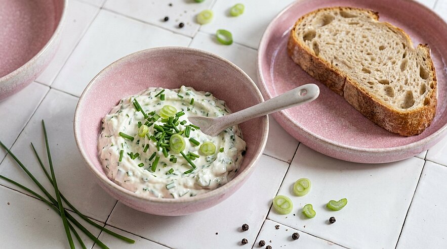 Cremiger Kräuteraufstrich mit gehacktem Schnittlauch und Frühlingszwiebeln in einer Schüssel mit Löffel; daneben eine Scheibe Bauernbrot auf einem Teller.