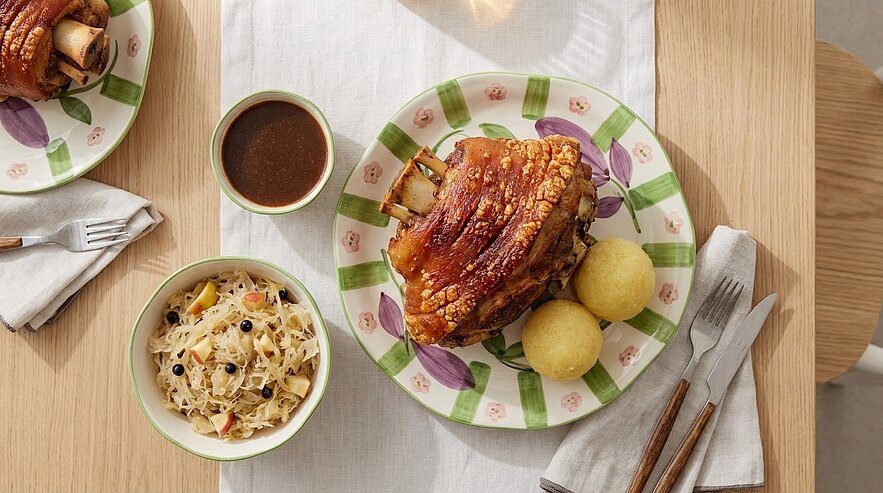 Knusprig gebratene Schweinshaxe mit zwei Kartoffelknödeln, Sauerkraut und brauner Bratensoße auf einem Teller.