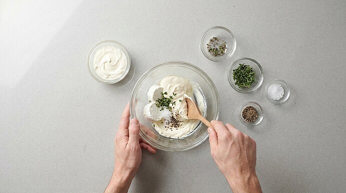 Zwei Hände verrühren Ziegenfrischkäse, Crème fraîche, Thymian, Salz und Pfeffer mit Holzlöffel in einer Glasschüssel.