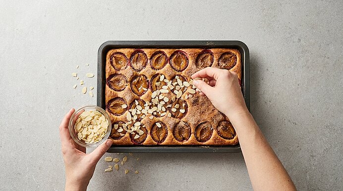 Zwei Hände streuen Mandelblättchen aus einer Schüssel auf einen frisch gebackenen Kuchen mit halbierten Pflaumen in einer rechteckigen Backform.