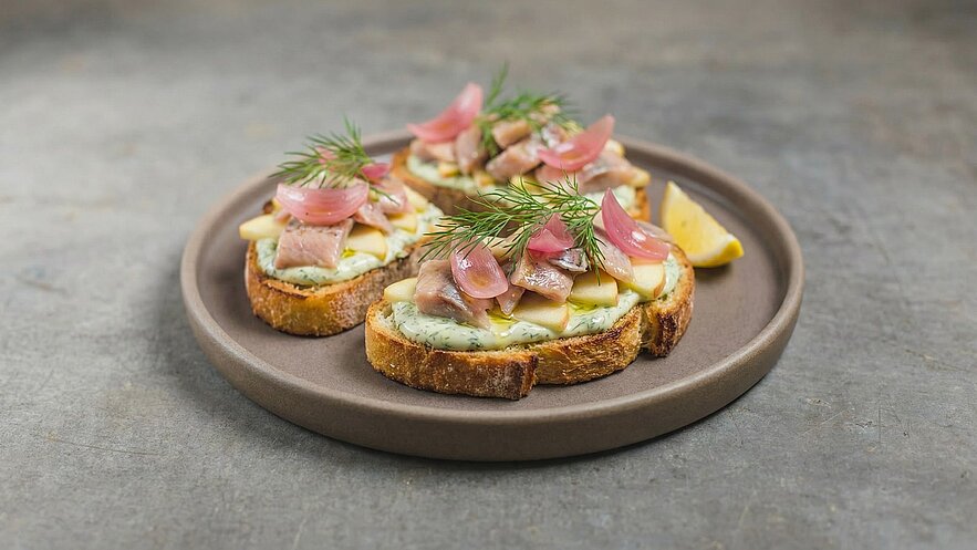 Drei kleine Brotscheiben liegen auf einem grauen Teller. Sie sind mit einer cremigen Sauce, Fischstücken, Äpfeln, roten Zwiebeln und frischem Dill garniert. Ein Stück Zitrone komplettiert die Präsentation.