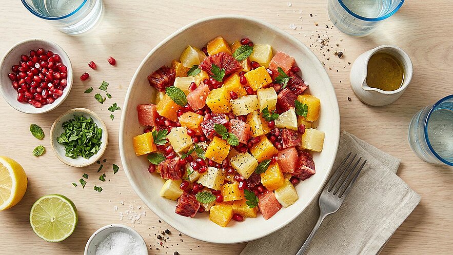 Eine große Schüssel mit frischem Obstsalat, bestehend aus bunten Stückchen von Melone, Ananas, Grapefruit und Granatapfelkernen. Der Salat ist mit frischen Minzblättern garniert und auf einem hellen Holztisch angerichtet.