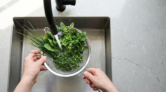 Zwei Hände halten ein Metallsieb mit frischen Kräutern (Schnittlauch, Petersilie) unter fließendem Wasser im Spülbecken zum Reinigen.