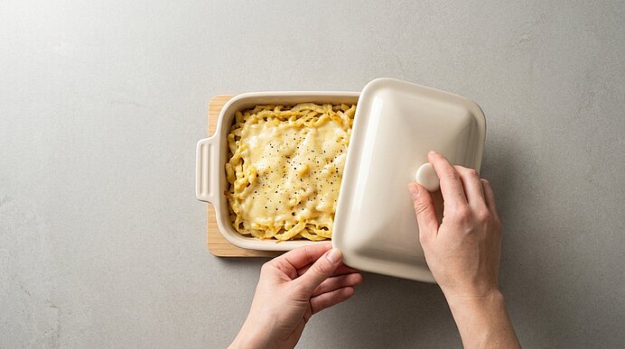 Zwei Hände heben den Deckel einer Auflaufform; darin mit Käse überbackene Bandnudeln, leicht mit schwarzem Pfeffer bestreut.