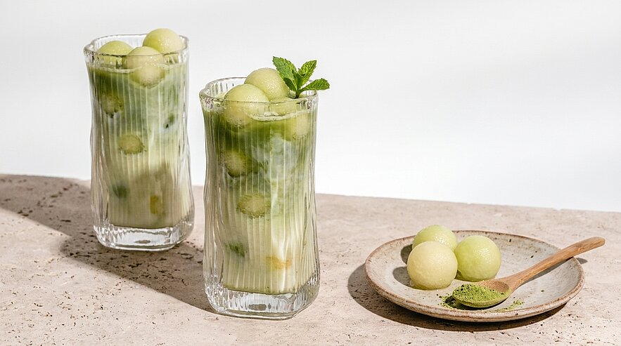 Zwei hohe Gläser mit grünen Melonenkugeln in milchigem Getränk, eines mit Minzblatt garniert; daneben kleiner Teller mit weiteren Melonenkugeln und Löffel mit grünem Pulver.