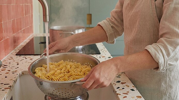 Person gießt Wasser über frisch gekochte Fusilli in einem Sieb im Spülbecken, um die Pasta abzuspülen.