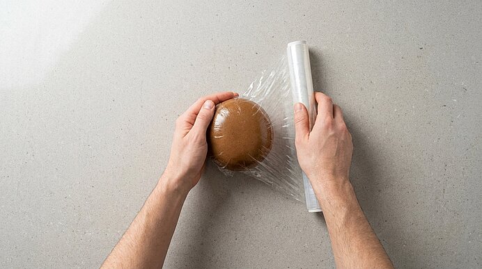 Zwei Hände wickeln ein rundes, braunes Gebäck mit transparenter Frischhaltefolie; eine Rolle Frischhaltefolie liegt daneben.