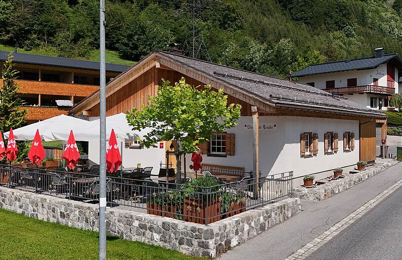 Ein rustikales Gasthaus mit einem Außenbereich, der von einem Geländer umgeben ist. Rote Sonnenschirme stehen auf der Terrasse, um Gästen Schatten zu spenden. Im Hintergrund sind grüne Hügel und weitere Häuser zu sehen.