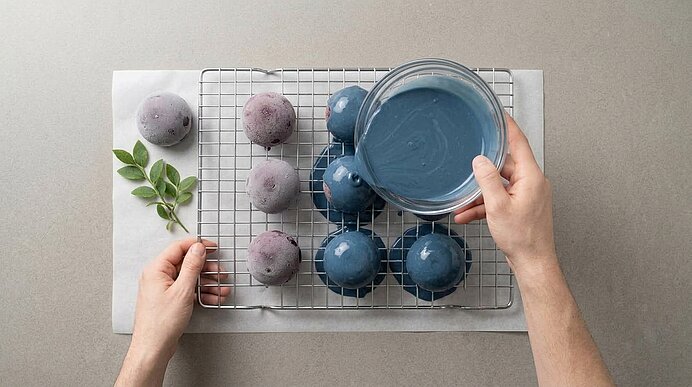 Zwei Hände gießen blaue Glanzglasur aus einer Glasschale über gefrorene Blaubeer-Dessertkugeln auf einem Kuchengitter.