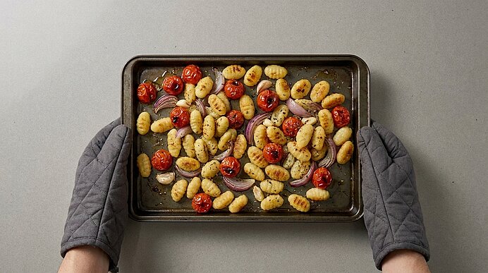 Backblech mit gerösteten Gnocchi, Kirschtomaten, roten Zwiebelringen und Kräutern, von zwei Händen mit Ofenhandschuhen gehalten.