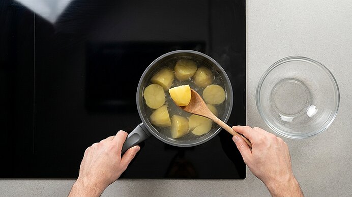 Draufsicht auf Hände, die mit einem Holzlöffel gekochte Kartoffelstücke in einem Topf auf dem Herd rühren; rechts steht eine leere Glasschüssel.